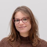 Sabrina La Rocca - Marketing Assistant: Smiling woman with red, round glasses and shoulder-length brown hair.
